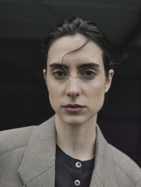 Portrait of a young woman with dark hair and brown eyes wearing a blazer against a dark background in a medium shot.