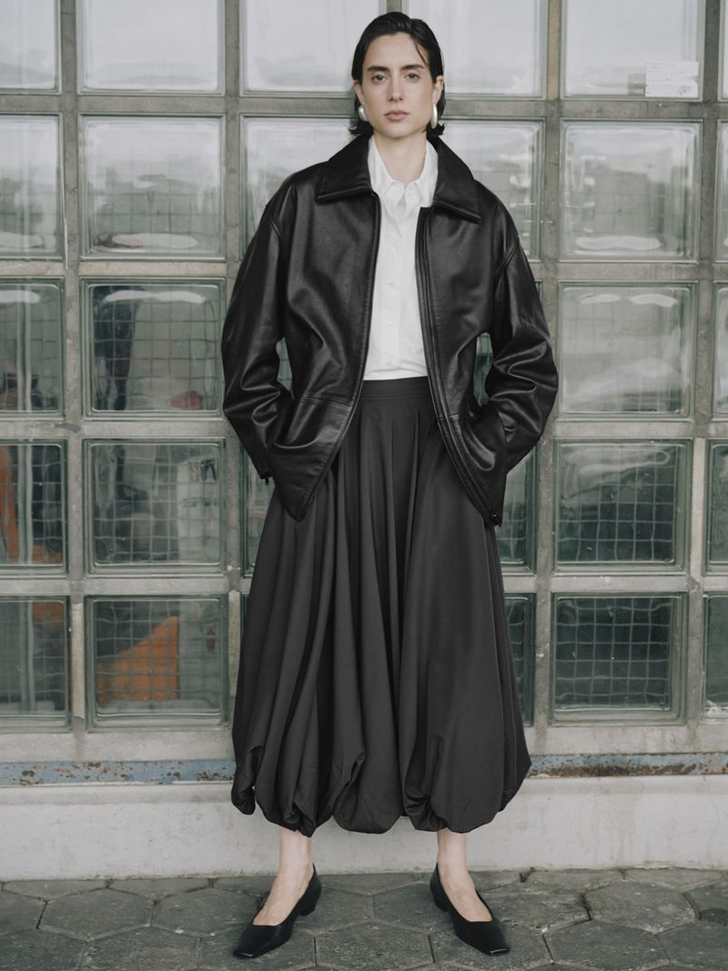 Woman in black leather jacket and voluminous skirt poses against a glass brick wall, creating a blend of modern fashion and urban style.