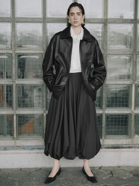Woman in black leather jacket and voluminous skirt poses against a glass brick wall, creating a blend of modern fashion and urban style.
