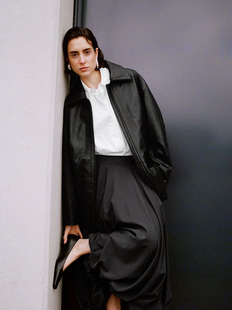 Portrait of a young adult female model with dark hair wearing a black leather jacket, white top, and dark pants leaning against a wall.