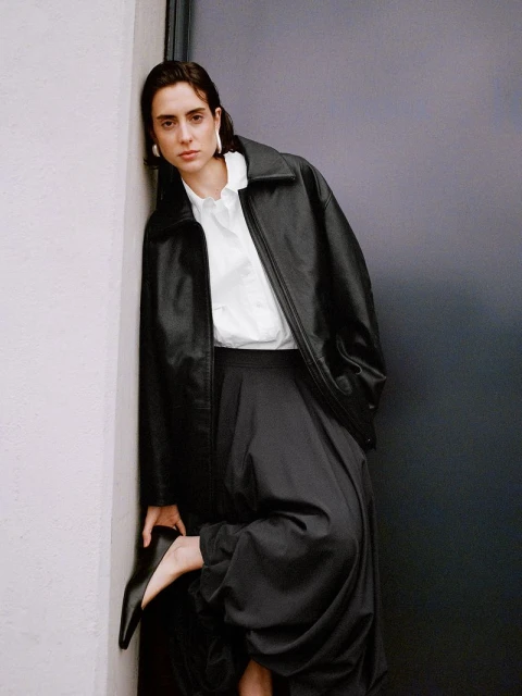 Portrait of a young adult female model with dark hair wearing a black leather jacket, white top, and dark pants leaning against a wall.