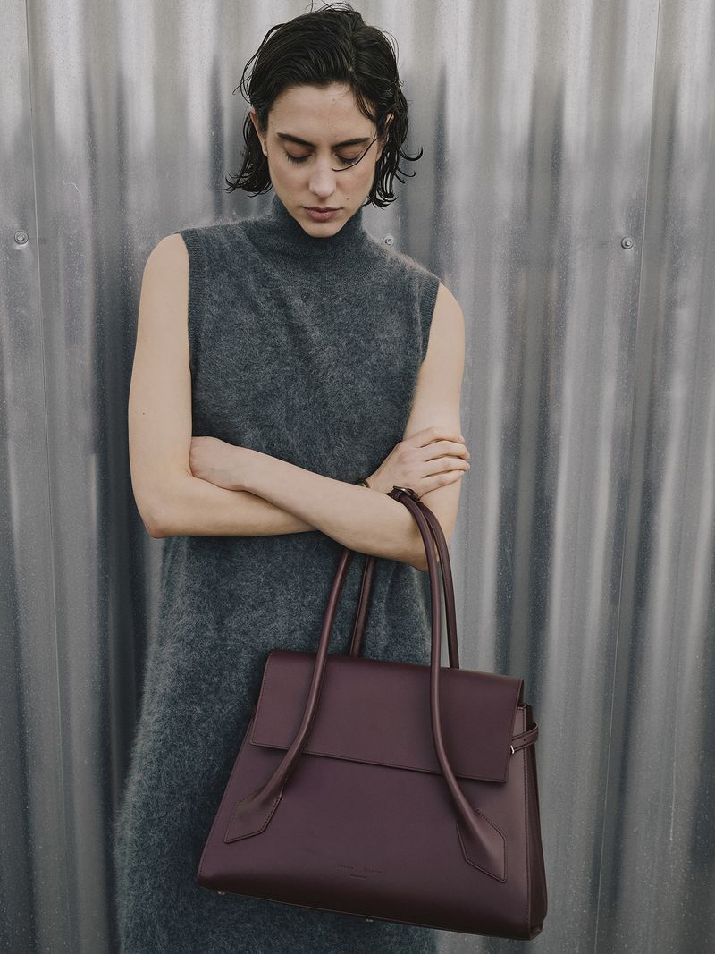 Brunette woman in grey dress holds a maroon leather handbag against a textured silver wall. Fashion shot, neutral colors, medium shot.
