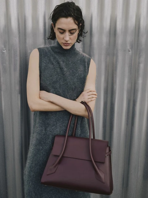 Brunette woman in grey dress holds a maroon leather handbag against a textured silver wall. Fashion shot, neutral colors, medium shot.