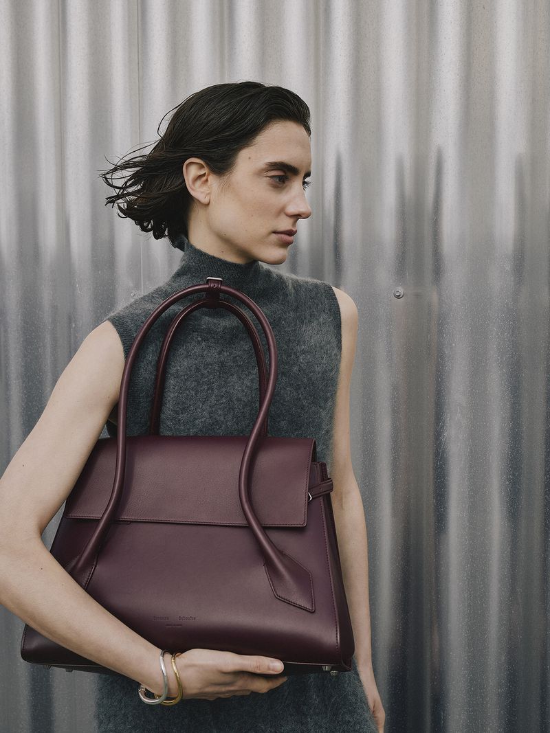A woman presents a modern dark red leather tote bag, accessorized with silver bracelets, against a textured metallic background.