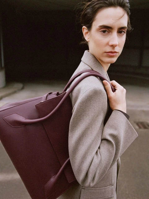 Woman in grey blazer carries a large purple leather tote bag looking directly at the camera in an outdoor urban setting.