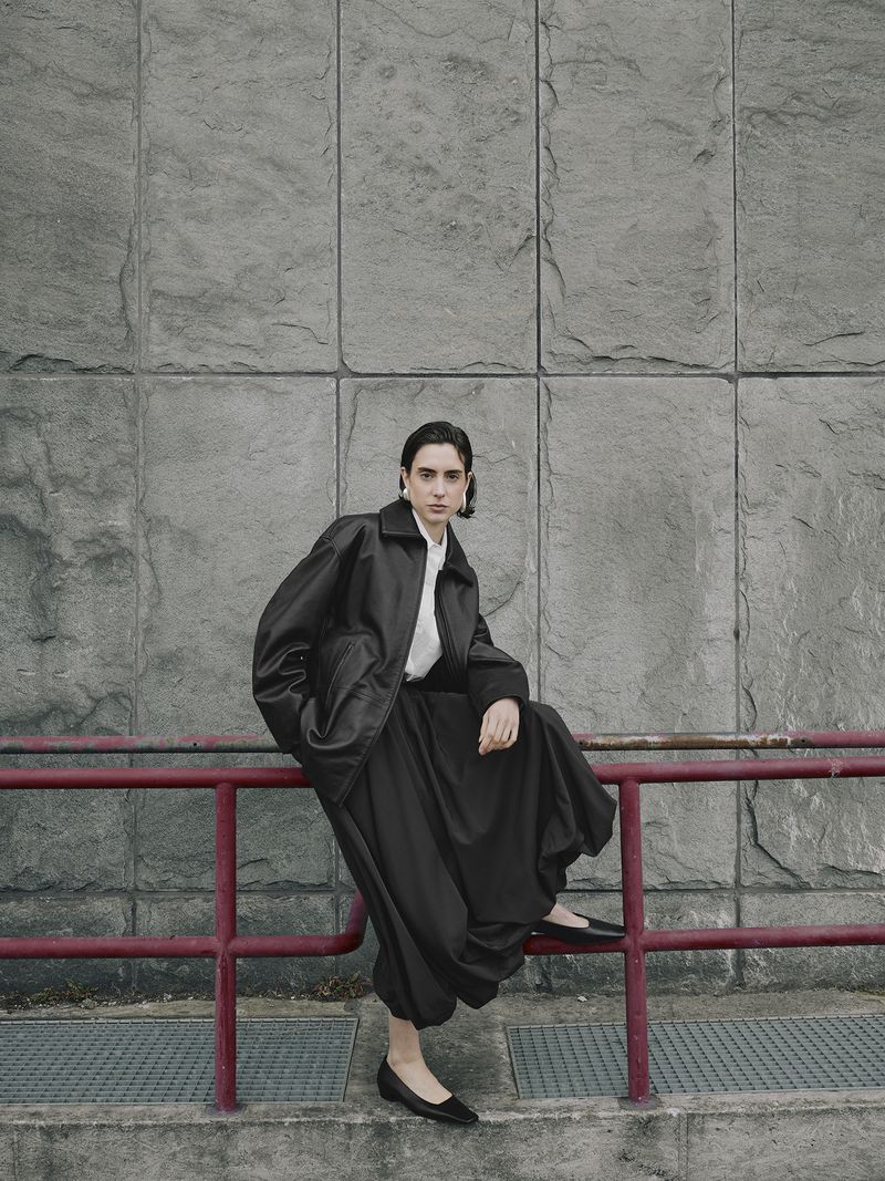 A stylish person in an oversized black jacket and pants sits on a red railing against a grey stone wall in an urban outdoor setting.