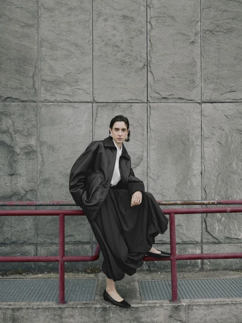 A stylish person in an oversized black jacket and pants sits on a red railing against a grey stone wall in an urban outdoor setting.