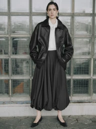 Beatriz Ronda stands wearing an oversized black leather jacket, white shirt, and voluminous black bubble skirt before a glass block wall.