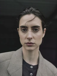 Close-up portrait of model Beatriz Ronda wearing a beige blazer over a dark buttoned top against a dark background