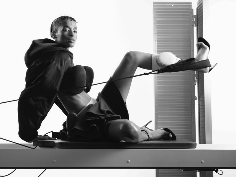 Athletic woman in black hoodie and shorts working out on pilates reformer machine in a brightly lit studio for fitness and wellness.