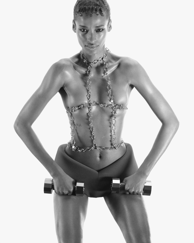 Athletic woman in sculptural chain top and workout shorts, holding dumbbells in a fitness-style black-and-white studio shot.