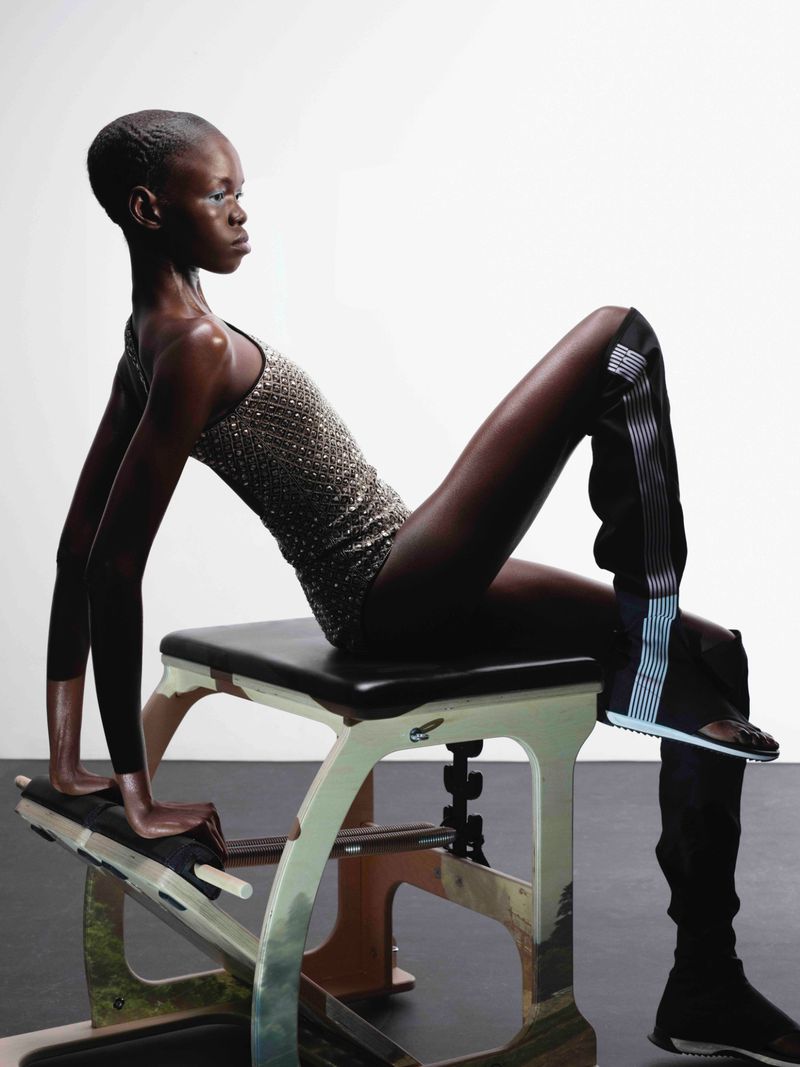 Elegant Black woman poses on a Pilates reformer machine in a photo studio, wearing a shimmering leotard and patterned leggings.