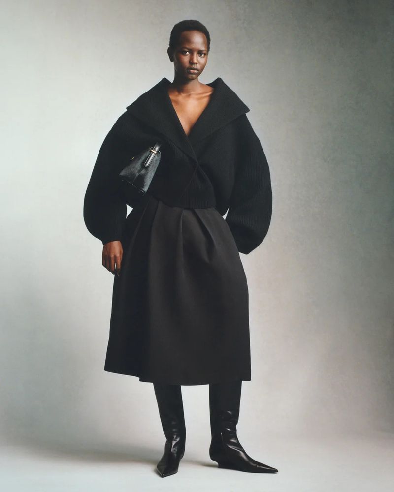 Fashionable Black model poses in all-black outfit: wrap jacket, A-line skirt, pointy boots. Bag in hand. Studio lighting, neutral background.