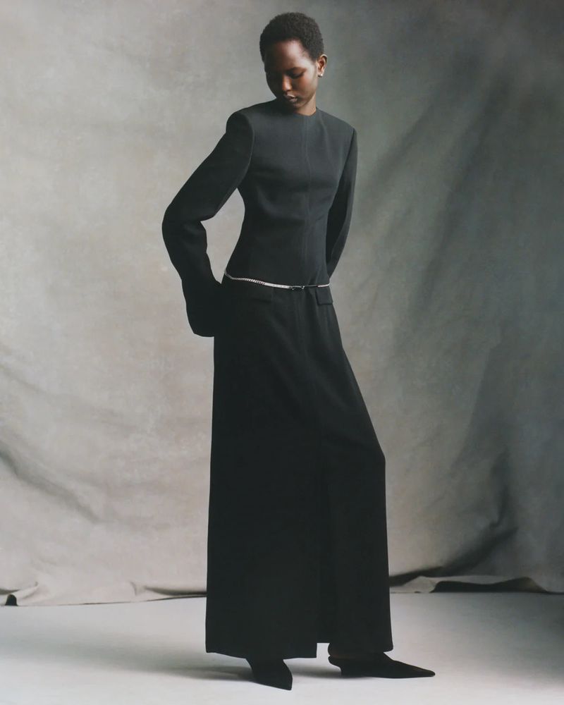 A woman stands in a full-length portrait in a dark dress, angled with her head turned toward her shoulder against a neutral backdrop.