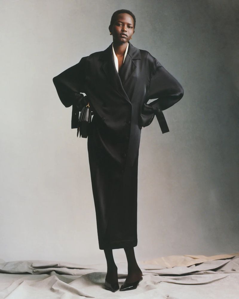 Fashionable adult woman in a black jacket and skirt, paired with black heels and a matching handbag, posing against a neutral backdrop.