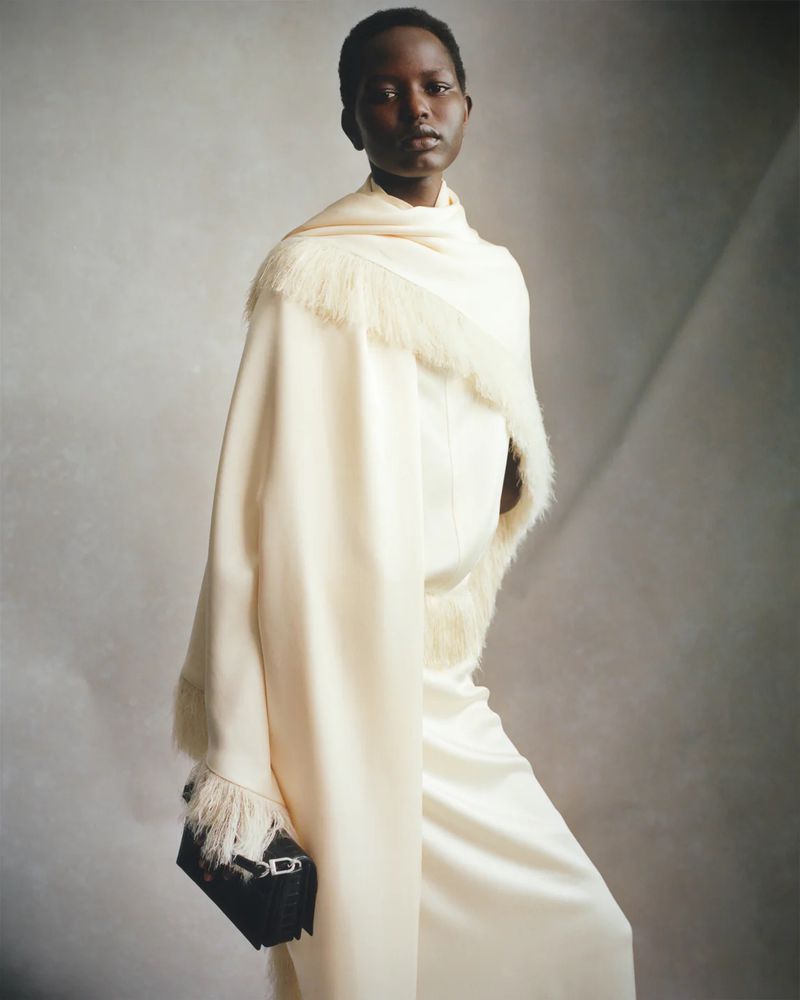 Elegant dark-skinned woman in white dress with shawl and black purse. Sophisticated fashion shot against neutral gradient backdrop.