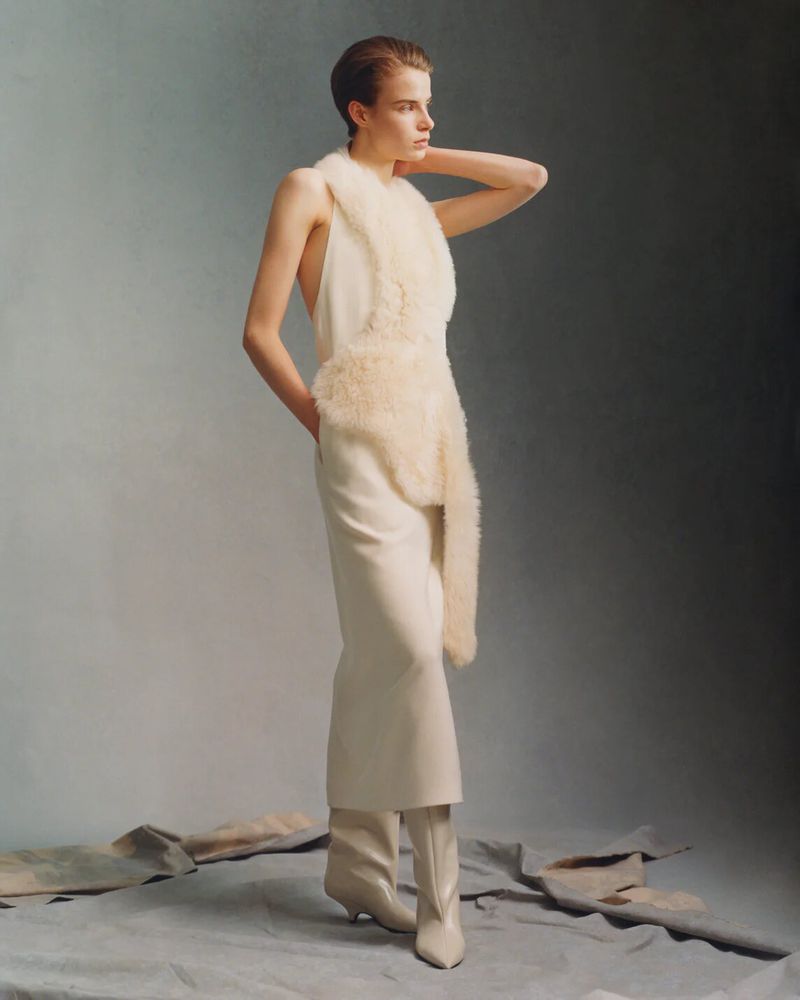 Model wearing a cream dress with textured trim and boots standing against a neutral studio backdrop for Toteme
