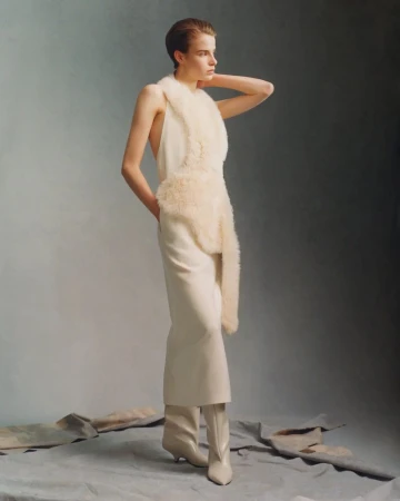 Model wearing a cream dress with textured trim and boots standing against a neutral studio backdrop for Toteme