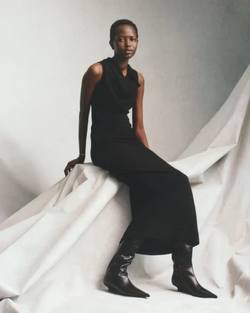 Elegant Black woman in a long dress and boots sits against a draped white fabric backdrop in a fashion editorial studio shot.