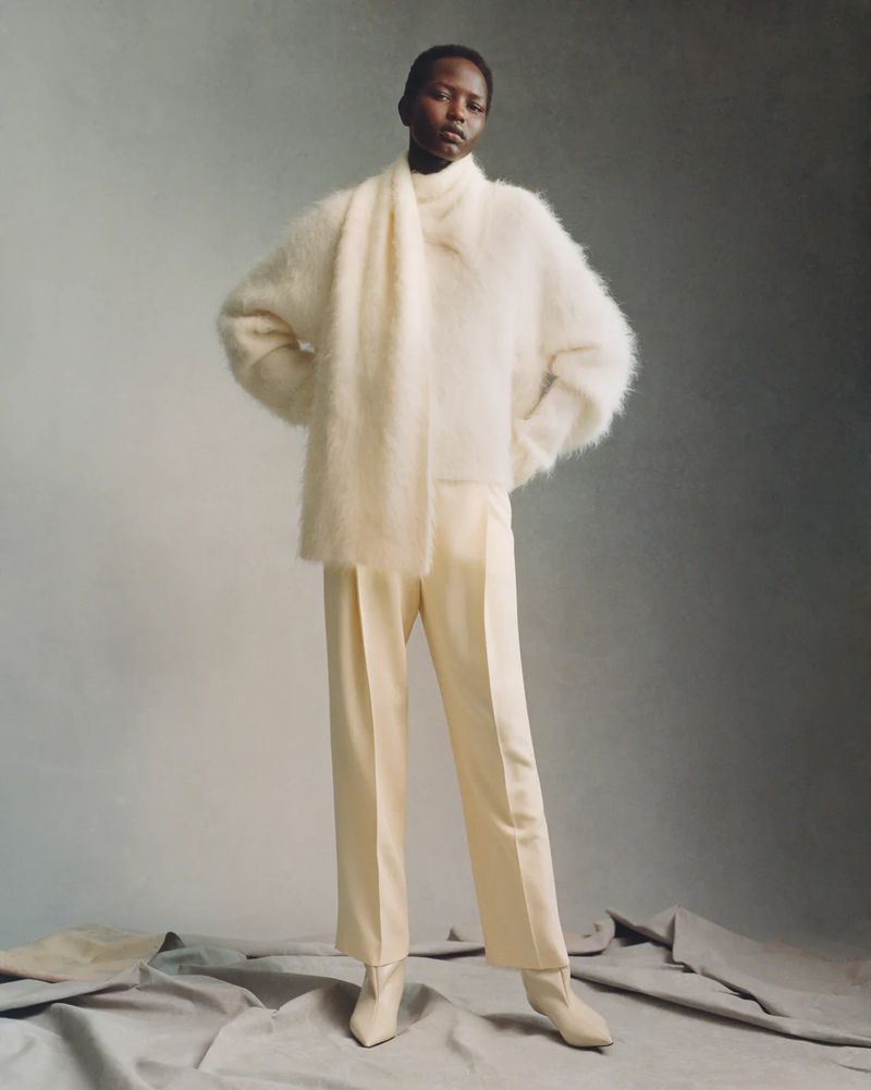 Elegant fashion shot of a black female model wearing a white sweater, scarf, pants, and heeled boots in a neutral-toned studio.