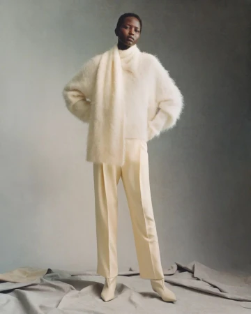 Elegant fashion shot of a black female model wearing a white sweater, scarf, pants, and heeled boots in a neutral-toned studio.