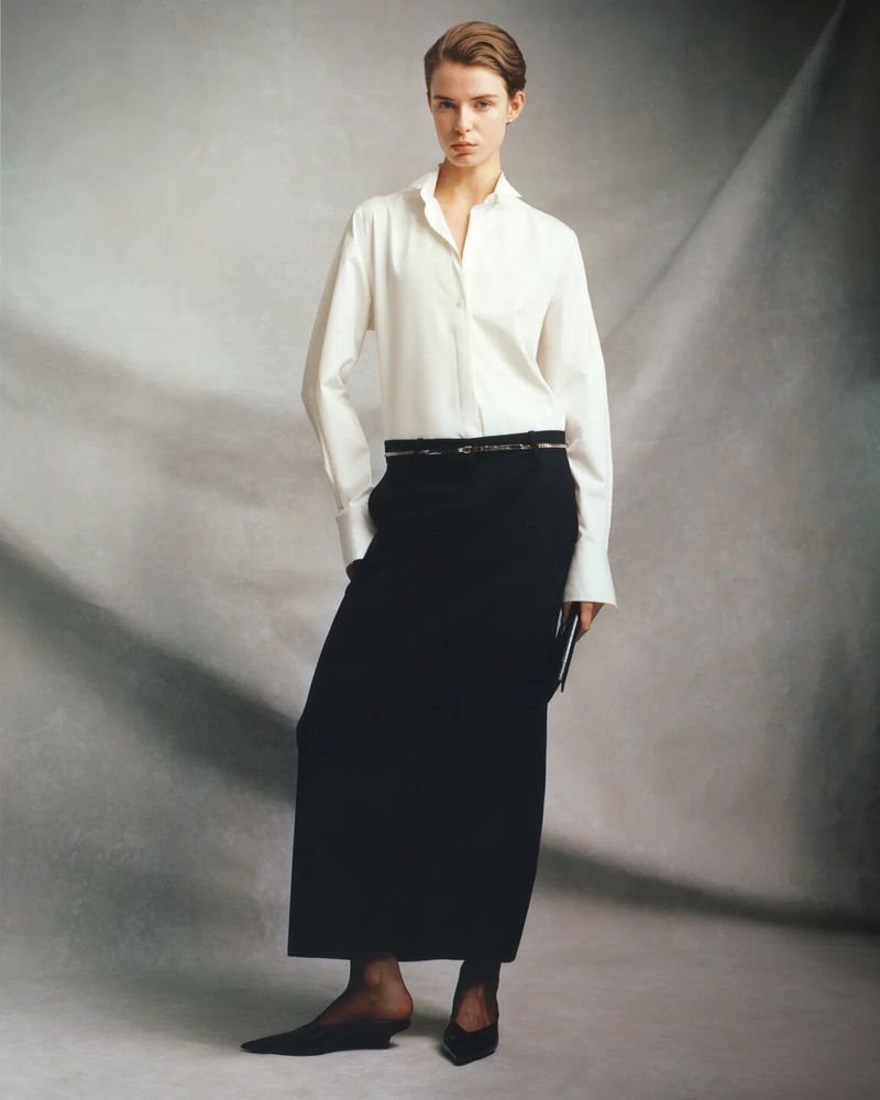 Full-length studio shot of a female model in a white shirt, long black skirt, and black pointed shoes, posing against a grey mottled background.