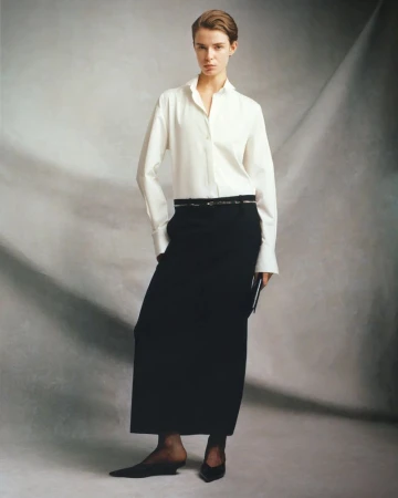 Full-length studio shot of a female model in a white shirt, long black skirt, and black pointed shoes, posing against a grey mottled background.