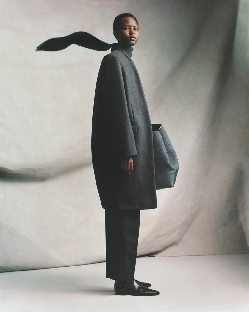 Fashion shot of Black woman wearing a large grey coat and black trousers, posing with a handbag on a cream-colored backdrop.