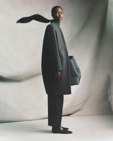 Fashion shot of Black woman wearing a large grey coat and black trousers, posing with a handbag on a cream-colored backdrop.