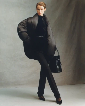 Fashion shot of a woman in a black oversized jacket, pants, and heels posing in a photo studio against a mottled grey backdrop.