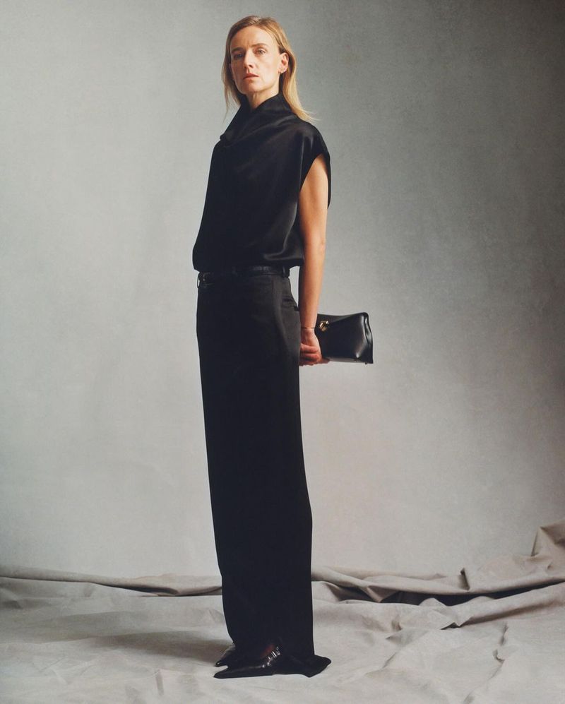 Adult woman posing in black top and black pants holding a black clutch in a studio setting. Full body shot with neutral background.