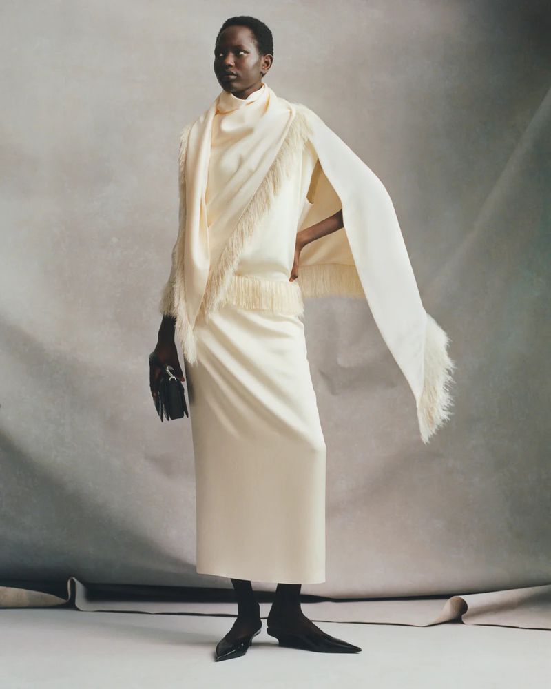 Elegant African woman in a cream dress and cape poses in a studio. She holds a black clutch and wears pointy-toe heeled shoes.