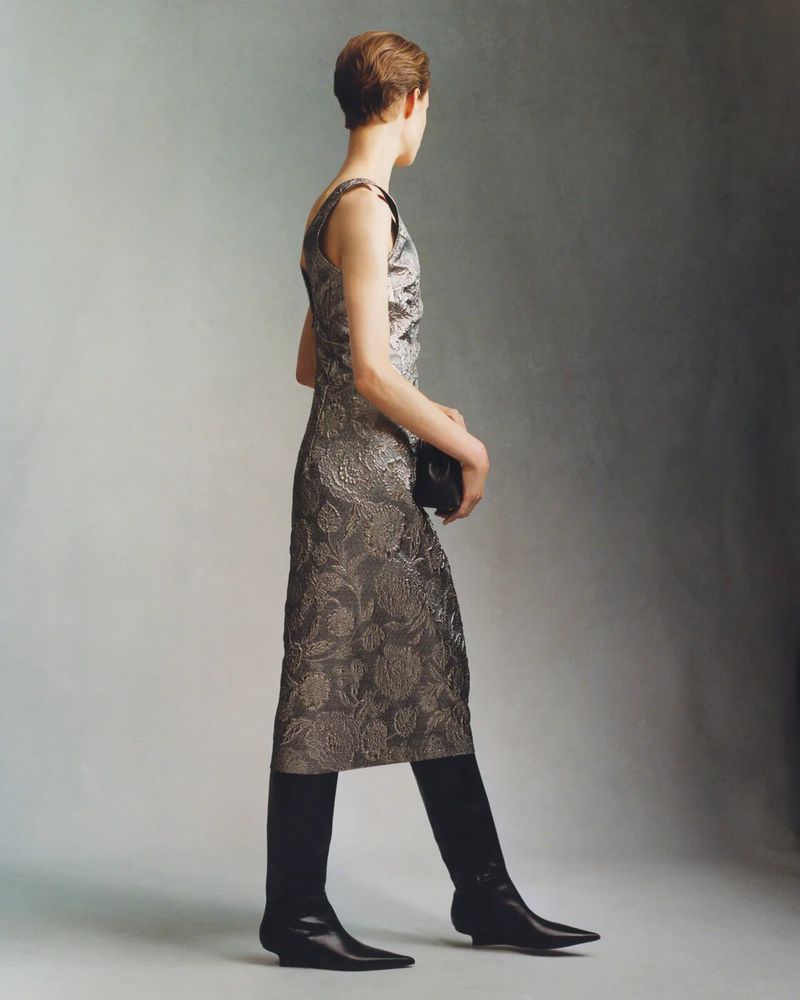 Model wearing a patterned metallic dress and black boots standing against a neutral grey studio background
