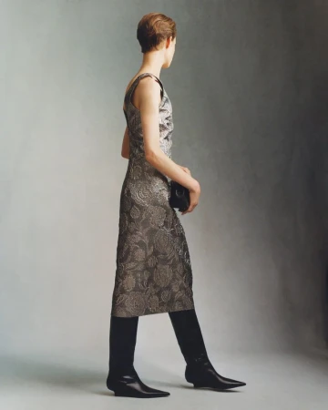 Model wearing a patterned metallic dress and black boots standing against a neutral grey studio background