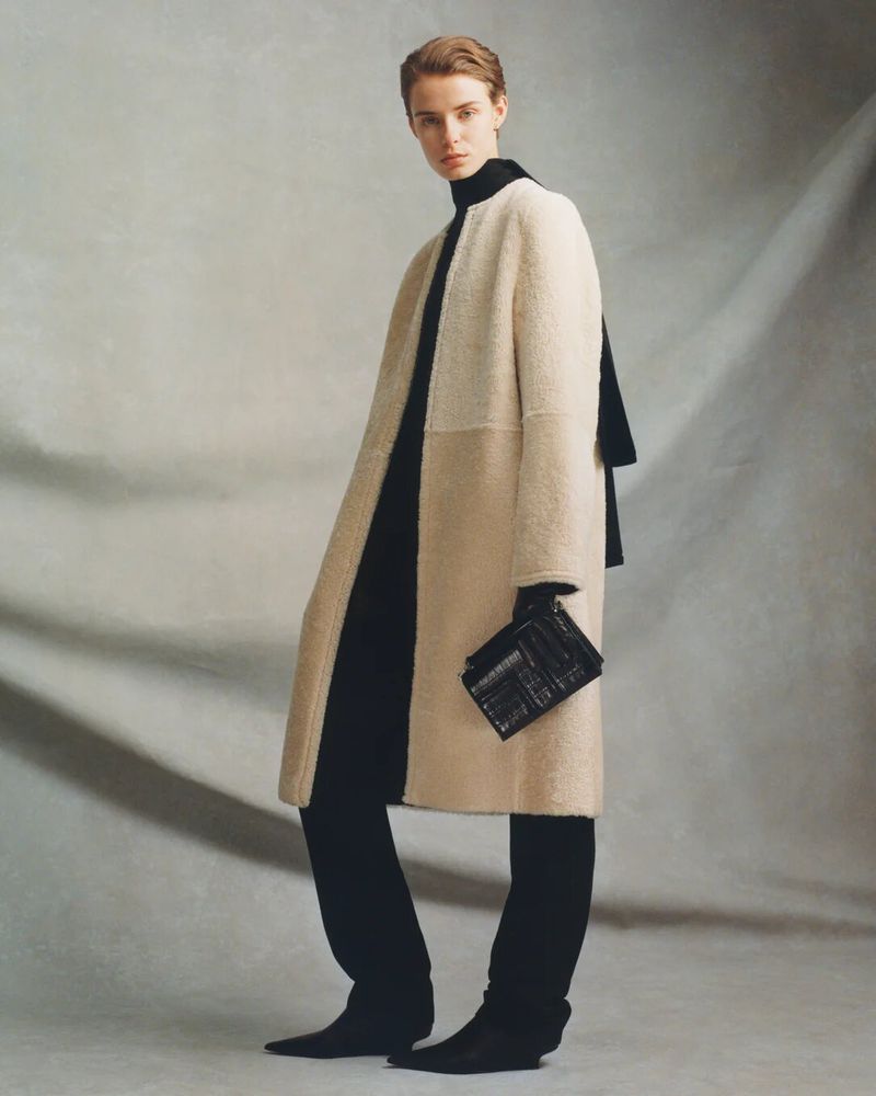 Fashionable adult woman posing indoors in a beige coat with a black purse, gloves, pants, and boots against a neutral backdrop.