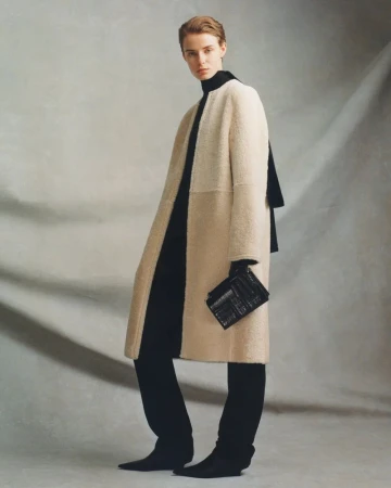 Fashionable adult woman posing indoors in a beige coat with a black purse, gloves, pants, and boots against a neutral backdrop.