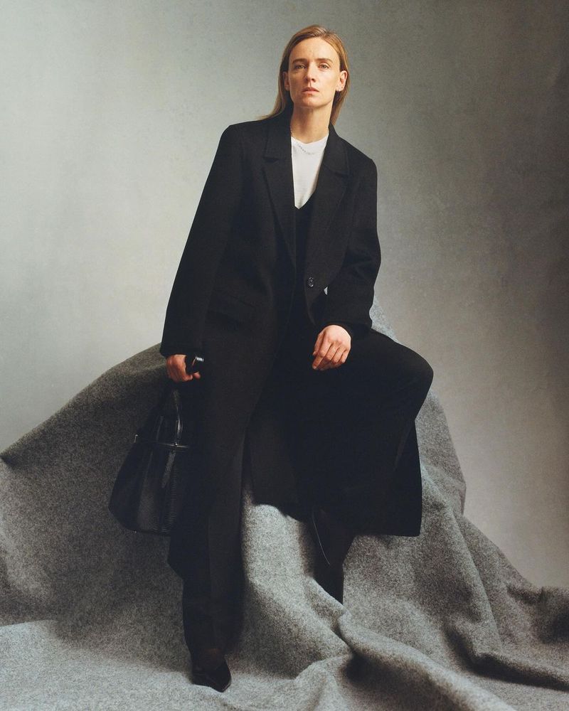A blond-haired woman in a black coat and pants sits on a gray textured surface in a fashion pose, set against a neutral gray background.