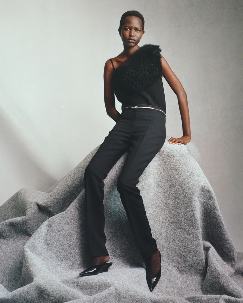 A fashion model with dark skin is seated on a gray fabric surface wearing a black top, black pants, and black heels in a studio shot.