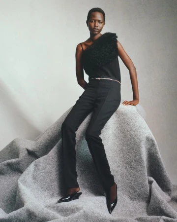 A fashion model with dark skin is seated on a gray fabric surface wearing a black top, black pants, and black heels in a studio shot.