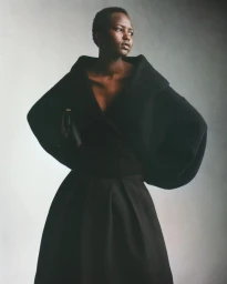 Model wearing a black knitted top and pleated skirt holding a handbag against a plain grey studio backdrop