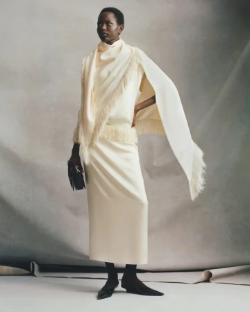 Model wearing a cream-colored fringe ensemble and cape holding a black clutch against a neutral seamless studio backdrop