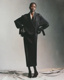 Model in black blazer and long skirt holding a handbag against a gradient backdrop
