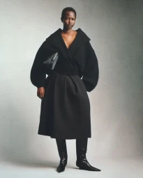 Model wearing a black sculptural jacket, skirt, and boots holding a handbag against a grey seamless backdrop