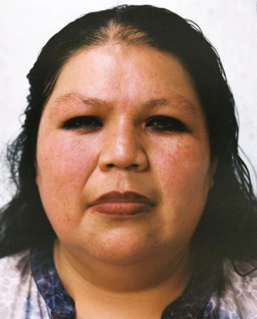 Close-up portrait of a woman with dark hair and dark eyeliner looking directly forward against a pale neutral background.