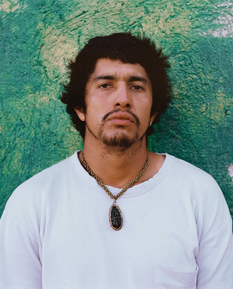 Portrait of a Hispanic man with dark hair and a goatee, wearing a white t-shirt and a large necklace, against a mottled green wall.