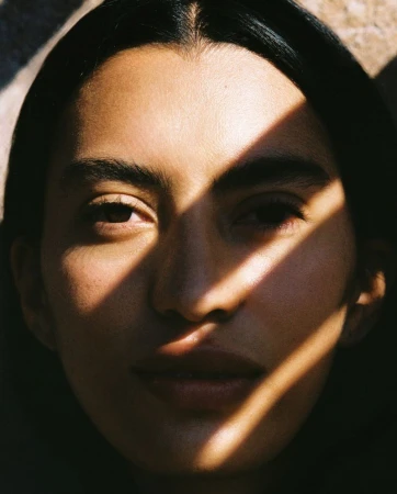 Close-up portrait of a woman with dark hair, partially illuminated by strong, directional sunlight creating sharp shadows across her face.