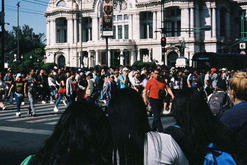 A large crowd of people walk across a street in front of an ornate building in Mexico City, with a bus visible in the background.