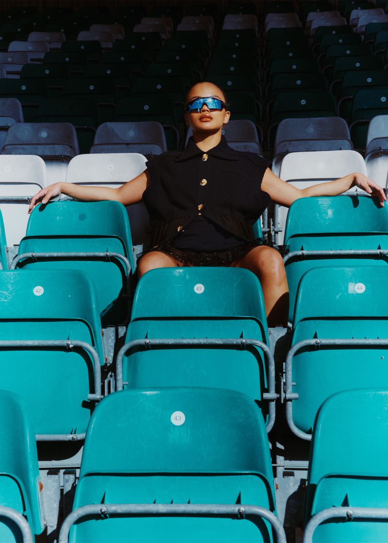 Stylish woman in sunglasses and a black top poses on an empty teal stadium seat, bathed in sunlight with visible shadows and textures.
