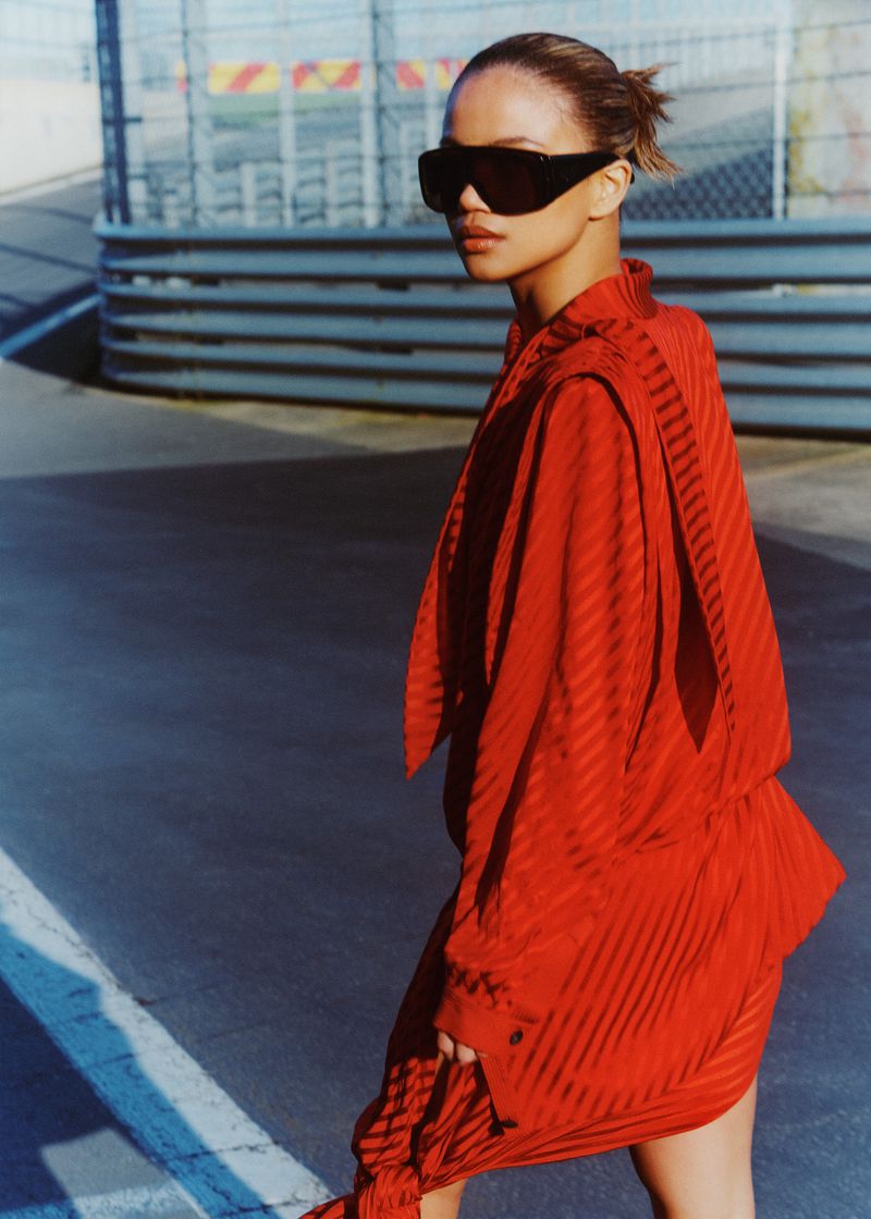 Woman in a red striped dress and sunglasses poses on a paved road with a concrete barrier in the background under bright sunlight.