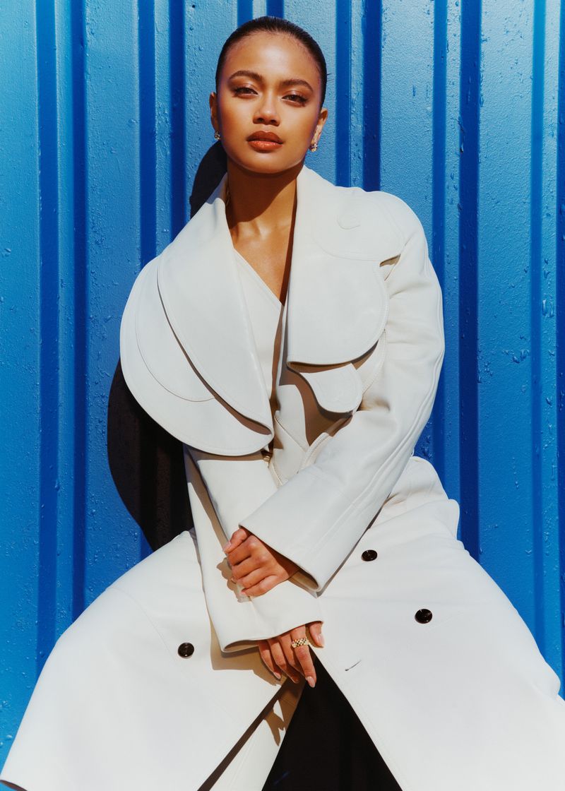 A woman in a white coat poses against a bright blue corrugated wall, hands clasped, in a fashion-style portrait with soft lighting.